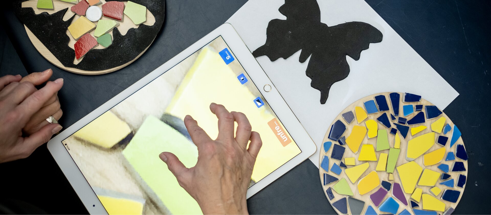 Hände arbeiten mit farbigen Mosaikteilen, einer schwarzen Schablone in Schmetterlingsform und einem Tablet auf einem Tisch.