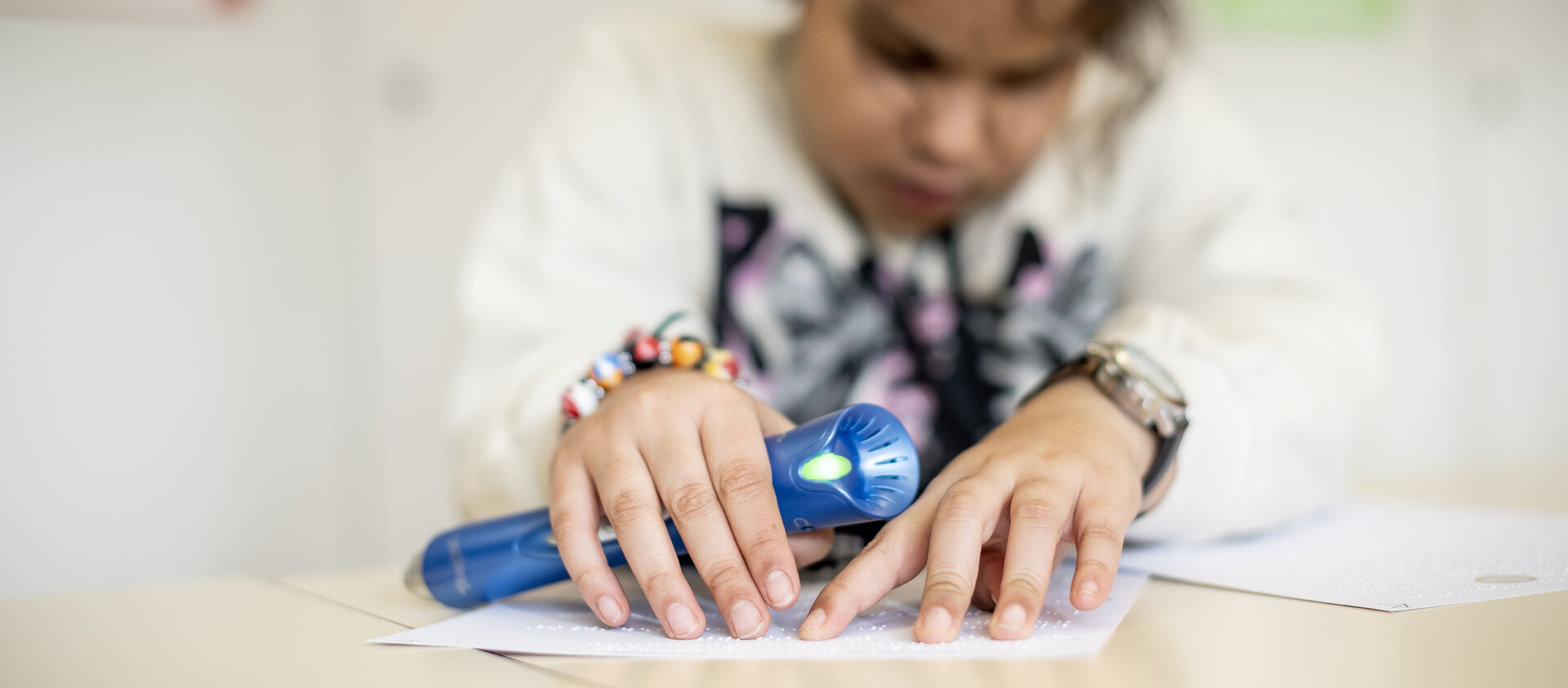 Ein Kind fährt mit den Fingern über Brailleschrift. In der Hand hält es ein blaues Lesegerät.