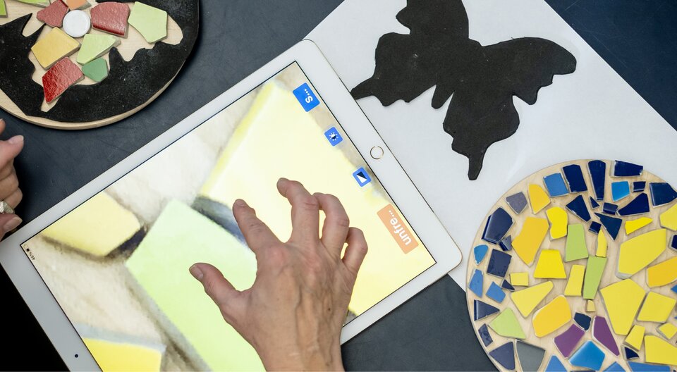 Hände arbeiten mit farbigen Mosaikteilen, einer schwarzen Schablone in Schmetterlingsform und einem Tablet auf einem Tisch.