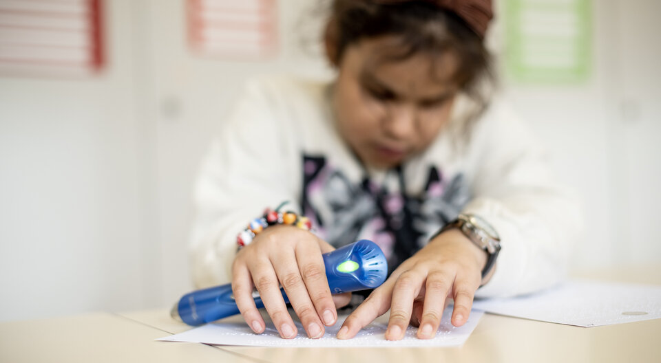 Ein Kind fährt mit den Fingern über Brailleschrift. In der Hand hält es ein blaues Lesegerät.